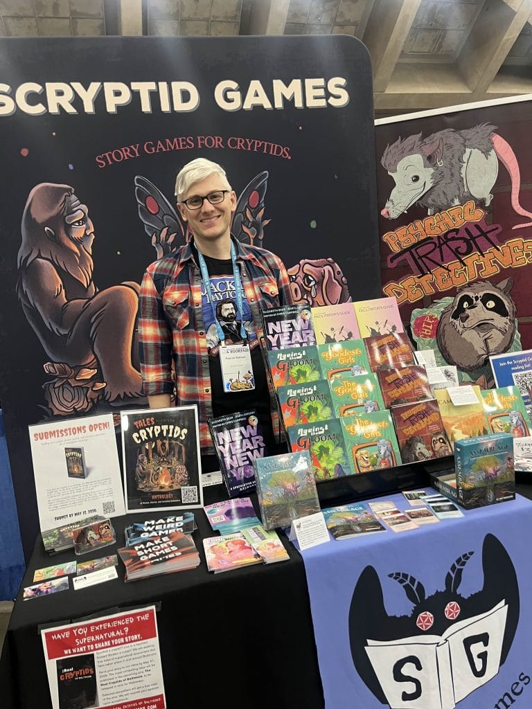 A photo of a man, Patrick Watson, standing at a booth for Scrpytid Games at a conference bookfair