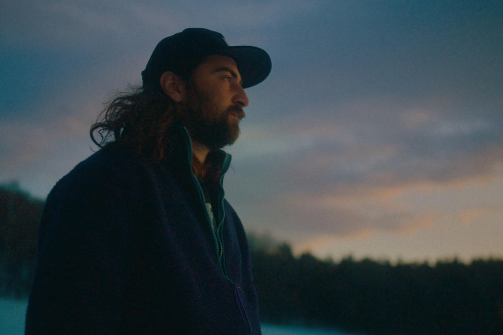A screenshot from the upcoming 2026 film Noah Kahan: Out of Body, showing a man standing in the woods in front of a sunset. 