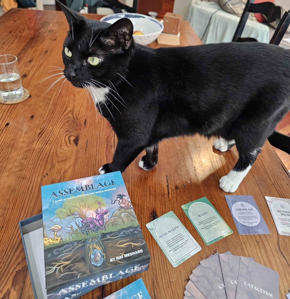 An image of a black-and-white cat walking on a wooden table next to the Assemblage TTRPG artwork and cards, featured in an Assemblage TTRPG review.