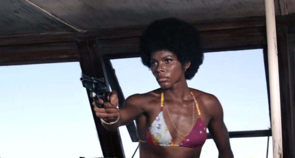 A screenshot from the 1973 James Bond movie Live and Let Die, showing Gloria Hendry portraying Bond girl Rosie Carver --- a Black woman with an afro wearing a bikini points a gun. This image is in an article about the character of Moneypenny in James Bond movies and video games.