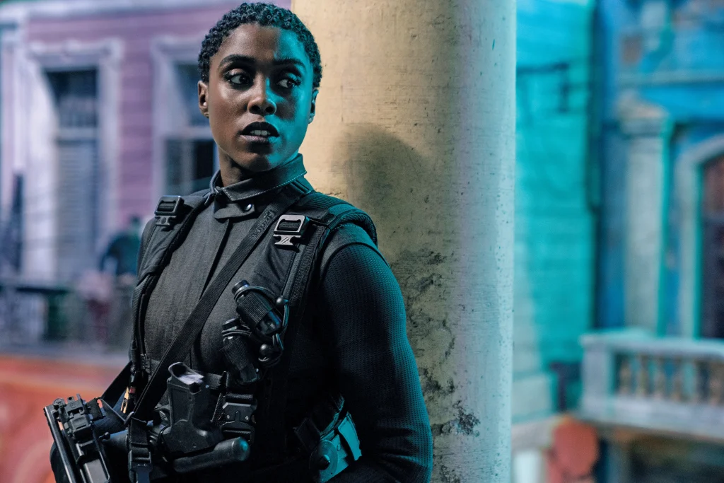 A screenshot from the 2021 James Bond movie No Time to Die, showing Lashana Lynch as Nomi, the new 007. A Black woman wears a black spy outfit with guns, leaning on a wall. This image is in an article about the character of Moneypenny in James Bond movies and video games.