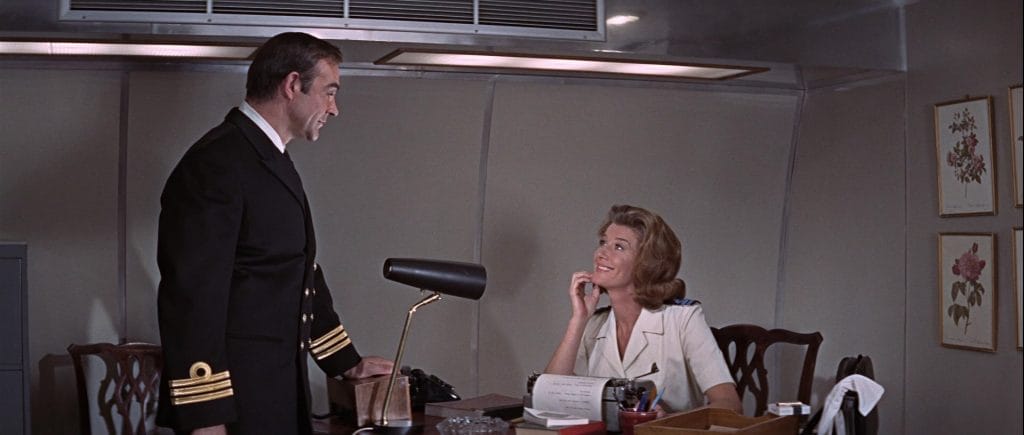 A screenshot from the film You Only Live Twice (1967) that shows a blonde woman in a naval uniform, Moneypenny, smiling up at Sean Connery as James Bond. This image is in an article about the character of Moneypenny in James Bond movies and video games.