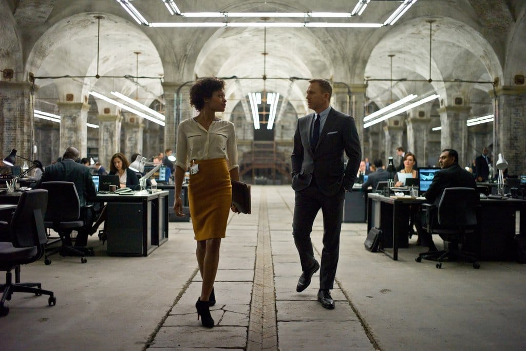 A screenshot from the 2012 James Bond movie Skyfall showing a modern looking underground computer lab and two people walking at the center: Moneypenny, portrayed by Black British actress Naomie Harris, and James Bond, portrayed by Daniel Craig. This image is in an article about the character of Moneypenny in James Bond movies and video games.